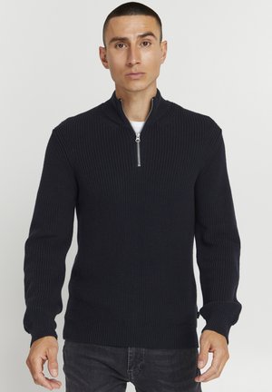 Marinblå stickad tröja med hög krage och halvmatch. Har ribbat mönster, långa ärmar och enfärgad design. Kombineras med mörka jeans.