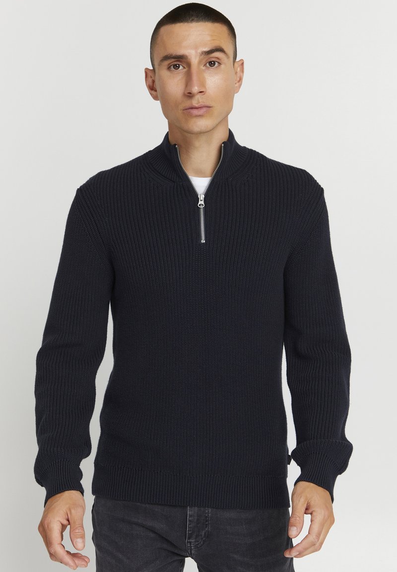 Marineblau gestrickter Pullover mit hohem Kragen und halbem Reißverschluss. Merkmale sind strukturierte Rippenoptik, lange Ärmel und einfarbig. Kombiniert mit dunklen Jeans.