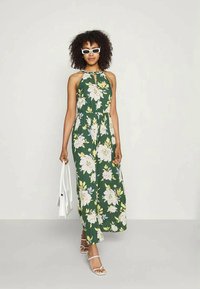 Mulher vestindo um vestido maxi halter floral verde, sandálias brancas e óculos de sol, segurando uma bolsa branca, em pé contra um fundo liso.