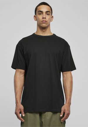 Junger Mann, der frontal steht und ein schlichtes schwarzes, übergroßes T-Shirt sowie olivgrüne Hosen vor neutralem Hintergrund trägt.