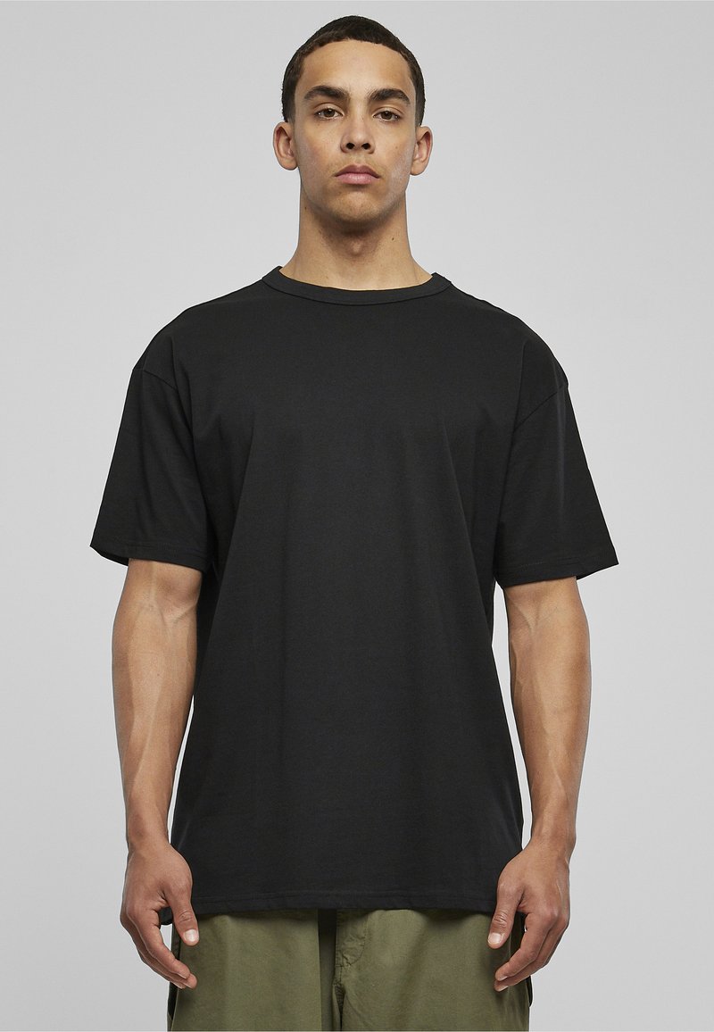 Jeune homme debout, face à l'avant, portant un t-shirt noir uni oversize et un pantalon vert olive sur un fond neutre.
