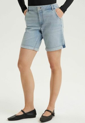 Osoba nosząca jasnoniebieskie krótkie spodenki jeansowe z podwiniętymi brzegami i czarną bluzkę z długim rękawem, w połączeniu z czarnymi balerinami Mary Jane.