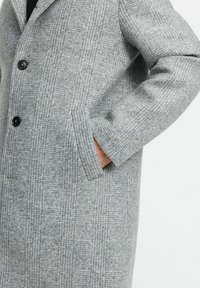 Hallhalline ruudulise villase mantli pikkade varrukatega, kahte eesmist taskut ja musti nuppe, millel on struktureeritud disain ja pehme tekstuur.