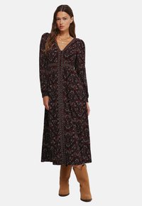 Dlouhé černé šaty s paisley vzorem v červené a modré barvě, s dlouhými rukávy, véčkovým výstřihem a centrálním knoflíkem. Spárováno s béžovými botami.