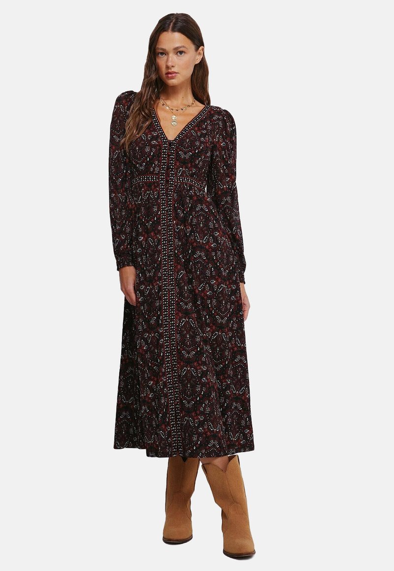 Dlouhé černé šaty s paisley vzorem v červené a modré barvě, s dlouhými rukávy, véčkovým výstřihem a centrálním knoflíkem. Spárováno s béžovými botami.