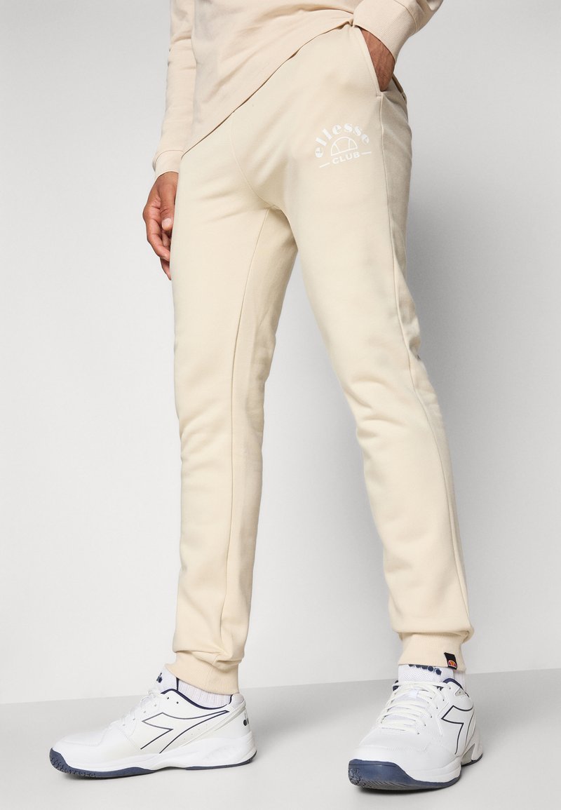 Beige Sweatpants aus weichem Material mit einem Logo auf der Vorderseite, schmal zulaufenden Beinen und gerippten Bündchen. Getragen mit weißen Sportschuhen.