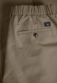 Pantaloni bej cu talie elastică, având o buzunar pe spate cu închidere prin buton și o etichetă mică de marcă aproape de buzunar.