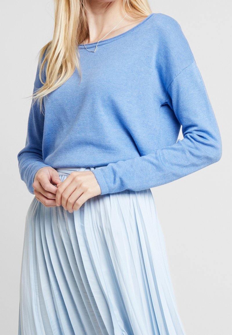 Ljusblå tröja i mjukt tyg, med vid halsringning och långa ärmar, i kombination med en veckad ljusblå kjol.