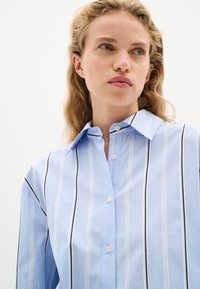 Camicia button-up azzurro chiaro con righe verticali bianche e blu scuro, dotata di collarino classico e una vestibilità comoda.
