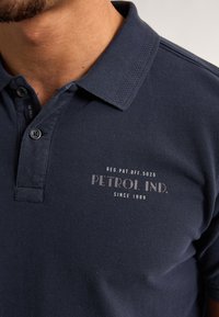 Námořnická modrá polo košile z texturovaného materiálu s límečkem, dvěma knoflíky a tištěným logem "PETROL IND." s doplňujícím textem níže.