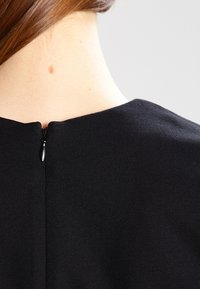 Czarny element odzieży o gładkiej teksturze, z tylnym zamkiem błyskawicznym i minimalistycznym designem. Dekolt znajduje się tuż nad ramionami.