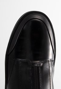 Black leather ankle boot with a smooth, glossy finish. Features a rounded toe and a side zipper for easy wear. Stitched accents on the upper.