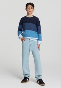 Niebieski sweter dzianinowy w poziome paski w ciemnoniebieskim i jasnoniebieskim odcieniu; zestawiony z jasnymi dżinsami i czarnymi sneakersami.