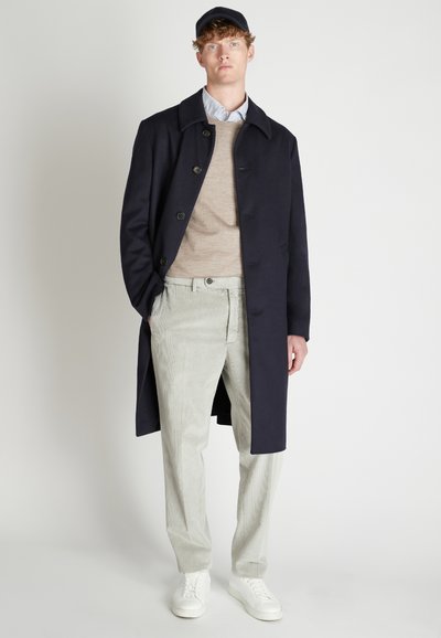 Marineblauer Woll-Trenchcoat mit großen Knöpfen, beigem Pullover, hellgrauen gestreiften Hosen und weißen Sneakern. Das Model steht vor einem schlichten Hintergrund.