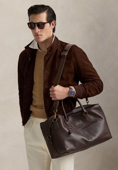 Homme portant une veste marron, un pull beige, un pantalon blanc, des lunettes de soleil et une montre tient un grand sac en cuir marron foncé avec une bandoulière.