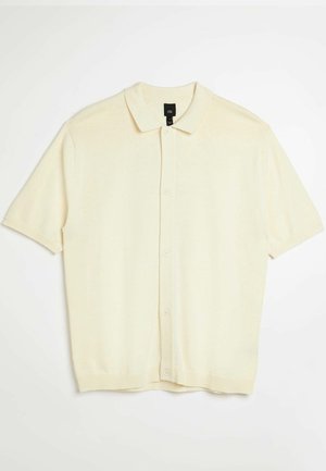 Polo a maniche corte di colore crema con una texture morbida, colletto classico e apertura frontale con bottoni. Presenta orlo e maniche a costine.