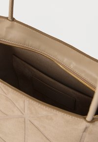 Beige színű táska strukturált kialakítással, sima bőr külsővel és texturált díszítésekkel. A belső térben cipzáras zseb és velúr bélelés található.