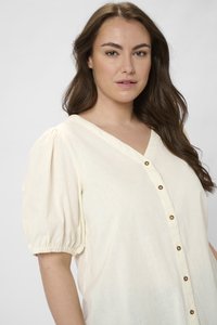 Blouse couleur crème avec des manches bouffantes, décolleté en V et boutons marron sur le devant. Fabriquée dans un tissu léger et texturé.
