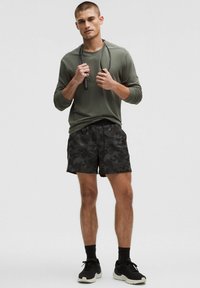 Maglietta sportiva a maniche lunghe verde oliva, pantaloni corti neri a motivo mimetico, scarpe da ginnastica nere. Il modello tiene una corda per saltare, mostrando un outfit casual per l'allenamento.