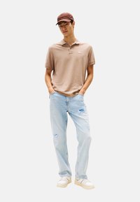 Polo marrón de manga corta, vaqueros desgastados azul claro y zapatillas de color claro, complementados con una gorra marrón. Ajuste y diseño casual.