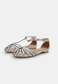 Sandales métalliques argentées avec un design découpé, semelle plate, bride de cheville et clous en forme de pyramide accentuant l'avant. Doublure intérieure lisse.