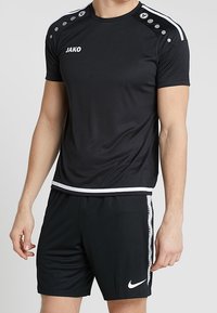 Černé sportovní tričko s šedými akcenty, které má tečkovaný design ramene. V kombinaci s černými šortkami, zdobenými bílým logem Nike.