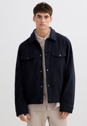 Joven vestido con una chaqueta negra abotonada sobre una camisa beige y pantalones, de pie contra un fondo claro y liso.