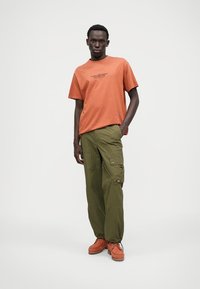 Tricou din bumbac portocaliu cu text imprimat, combinat cu pantaloni cargo de culoare verde măslin, cu buzunare, și încălțăminte portocalie cu tălpi texturate.