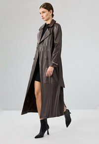 Bruine leren trenchcoat met een dubbelzijdig ontwerp, grote kraag, zijsplitten en accenten van gouden knopen, gedragen over een zwarte jurk.