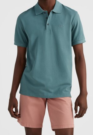 Uomo con polo a maniche corte color turchese e pantaloncini rosa chiaro, in piedi con le braccia rilassate davanti a uno sfondo semplice.