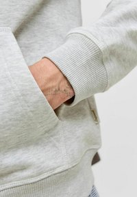 Hellgrau Sweatshirt mit strukturiertem Finish, ausgestattet mit gerippten Bündchen und einer frontalen Tasche. Die Hand befindet sich in der Tasche und zeigt ein Tattoo-Detail.