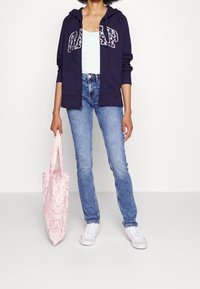 Felpa con zip blu navy con lettering floreale "GAP", top leggero blu chiaro, jeans blu chiaro e borsa a tracolla rosa con motivo, abbinati a sneakers bianche.