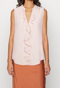 Blouse rose pâle sans manches avec un devant à volants, un décolleté en V, un tissu doux et un ourlet arrondi, associée à une jupe couleur rouille.