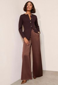 Brauner Strickcardigan mit rundem Ausschnitt und goldenen Knöpfen, kombiniert mit beigefarbenen, weiten Satin-Hosen und spitzen braunen Pumps.
