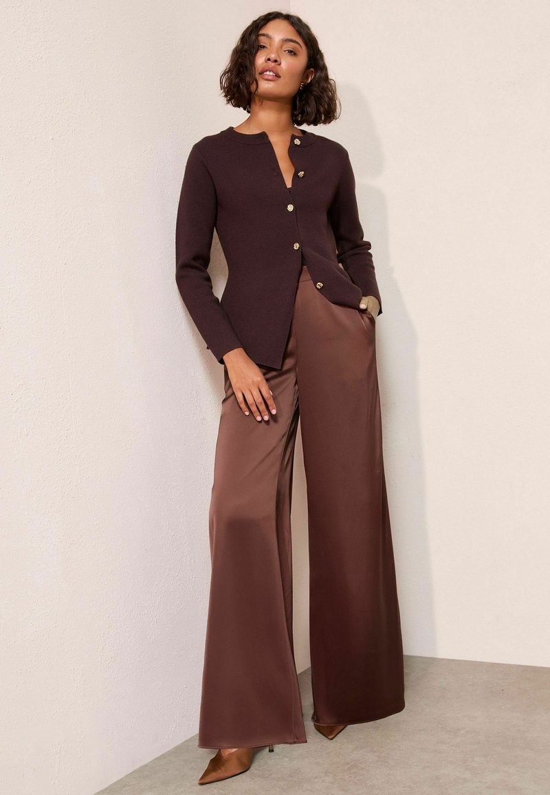 Brauner Strickcardigan mit rundem Ausschnitt und goldenen Knöpfen, kombiniert mit beigefarbenen, weiten Satin-Hosen und spitzen braunen Pumps.