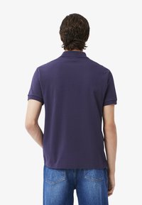 Man i en lila pikétröja, sedd bakifrån, med korta ärmar och denimjeans. Enkel vit bakgrund.