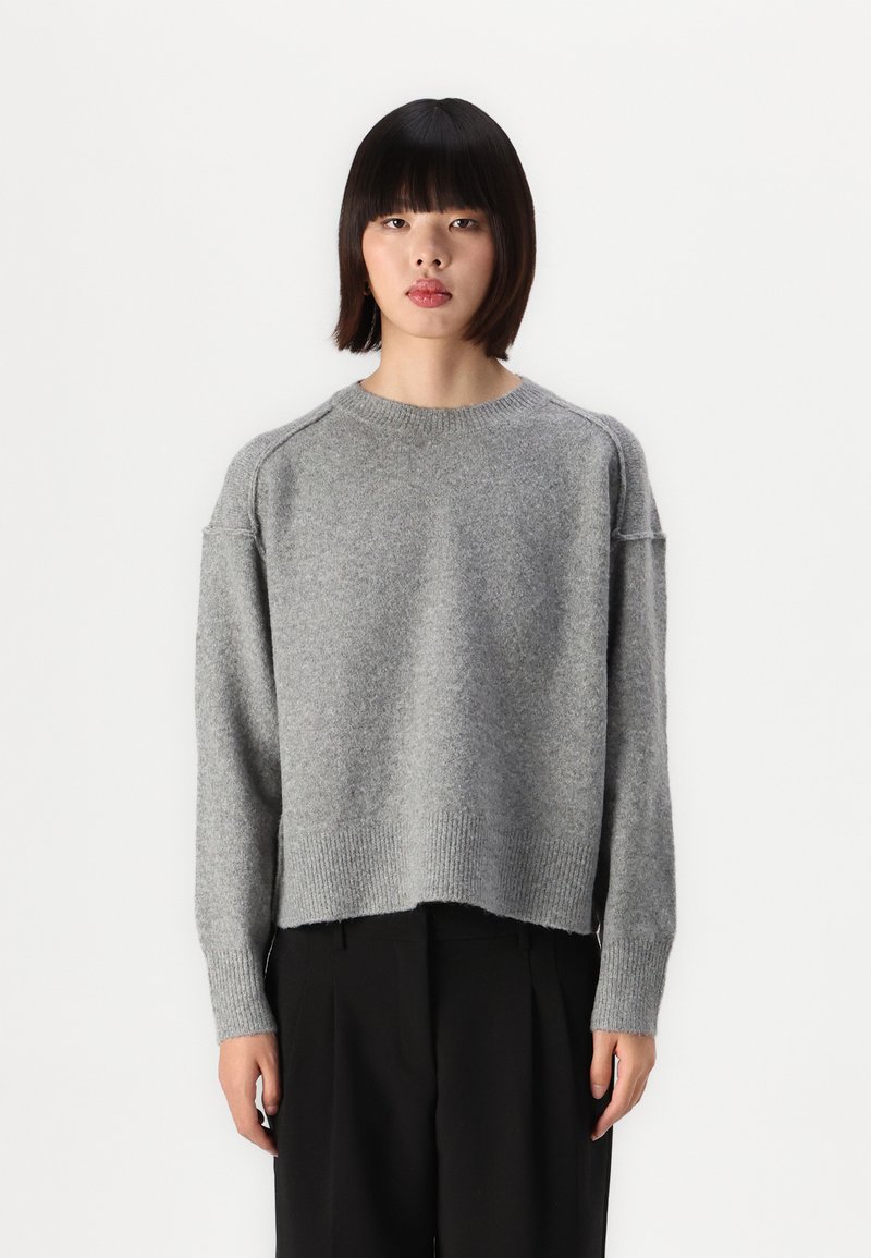 Grå strikkegenser med rund hals, senkede skuldre og ribbestrikket kant. Stoffet ser mykt ut med en litt fuzzy tekstur.