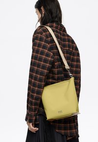 Persona che indossa una camicia a quadri nera e arancione, con una borsa a tracolla Bimba y Lola di colore verde chiaro e tracolla beige, vista da dietro.