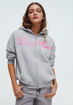 Junge Frau trägt eine hellgraue Zip-up-Hoodie mit pinker Aufschrift "Loud Noises", die Hände in den Taschen und passende graue Jeans vor weißem Hintergrund.