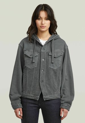 Junge Frau mit dunklen Haaren, die eine graue Jeansjacke mit Kapuze und dunkle Hosen trägt, steht vor einem schlichten hellen Hintergrund.