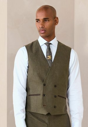 Man wearing olive green double-breasted vest over white dress shirt with patterned tie and gold lion tie clip, standing against beige background.