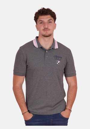 Homme portant un polo gris avec des accents roses et un jean bleu, debout les mains dans les poches devant un fond uni.