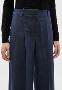 Persona, kas valkā tumši zilas, ar augstu jostasvietu un platām kāju daļām bikses, un melnu garām piedurknēm džemperi, rokas atslābinātas gar sāniem, vienai rokai uz delnas mazs tetovējums.