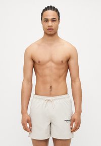 Beige svømmeshorts med snøre i livet og sidespalter, med en sort logo. Modellen har kort flettet hår og en veltrent fysikk.