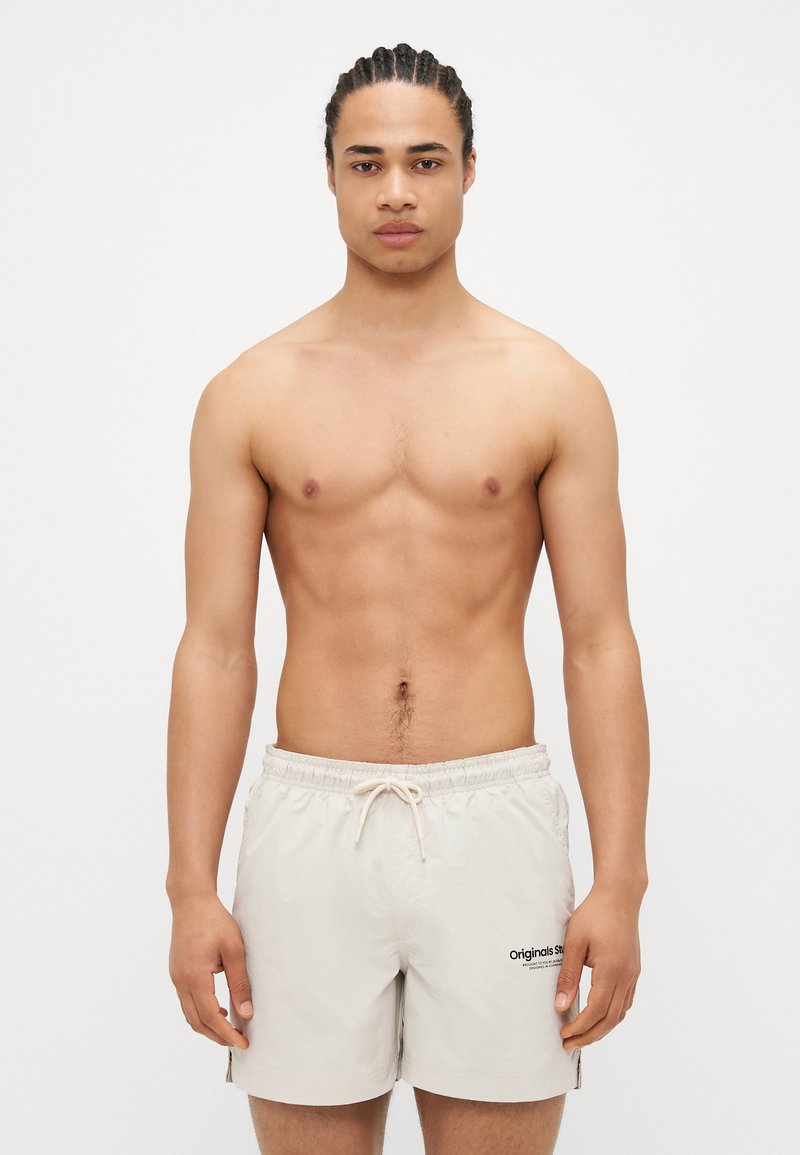 Beige svømmeshorts med snøre i livet og sidespalter, med en sort logo. Modellen har kort flettet hår og en veltrent fysikk.