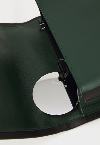 Sac en cuir vert avec une finition brillante, présentant un intérieur noir et un miroir rond en argent avec "PIERRE HARDY" gravé dessus.