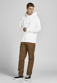 Witte hoodie met trekkoorden, gecombineerd met bruine broeken. Zwarte sneakers met witte veters en een eenvoudig ontwerp.