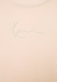 Ljusbeige t-shirt med en mjuk struktur, med en broderad signatur i ett kontrasterande off-white tråd nära kragen.