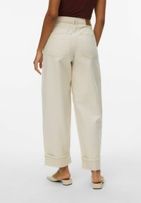 Personne portant un pantalon crème taille haute à jambes larges avec revers roulés et des chaussures beige à talons bas sans lacets, debout devant un fond blanc.