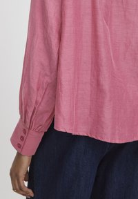 Blusa rosa leggera con maniche lunghe a sbuffo e polsini con bottoni. L'orlo scende leggermente sotto la vita, con una texture liscia.